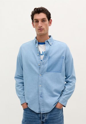 Light blue button-up shirt made of denim, featuring a single front pocket, long sleeves, and a relaxed fit. Worn over a white t-shirt.
