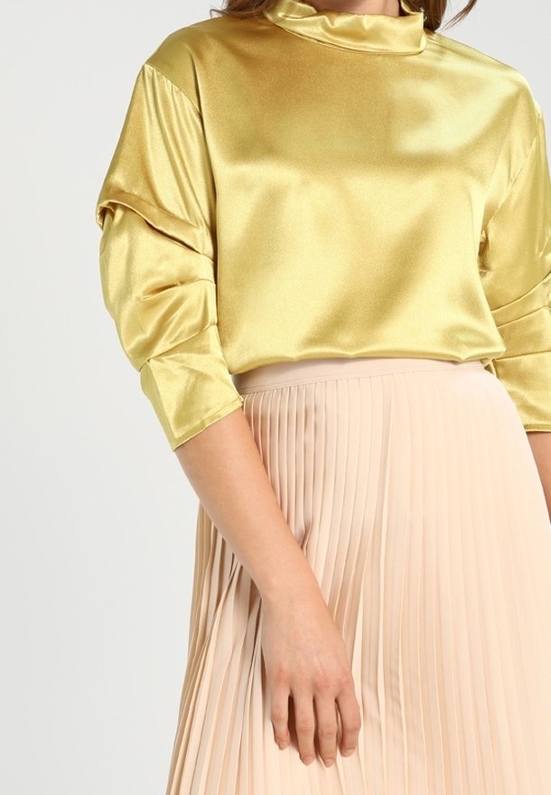 Femme portant un haut doré brillant à manches longues avec des manches froncées et un col montant, associé à une jupe plissée beige.