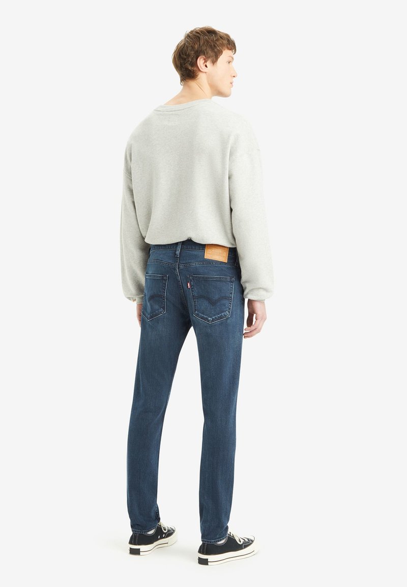Lichtgrauer Sweatshirt, Rundhalsausschnitt, kombiniert mit dunkelblauen Jeans mit braunem Lederpatch und klassischem Fünf-Taschen-Design. Schwarze Sneakers.