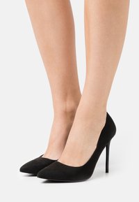 Černé semišové vysoké podpatky s designem špičaté špičky. Mají štíhlý stiletto podpatek a hladký povrch, což zdůrazňuje minimalistickou eleganci.
