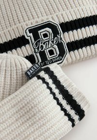 Strickmütze in hellbeige mit strukturiertem Rippenmuster, versehen mit schwarzen Streifen und einem gestickten "B"-Logo-Patch in Schwarz und Weiß.