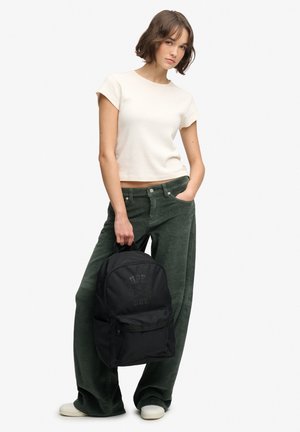 Mujer joven con camiseta blanca de manga corta y pantalones verdes holgados, sosteniendo una mochila negra con una mano, de pie sobre fondo blanco.