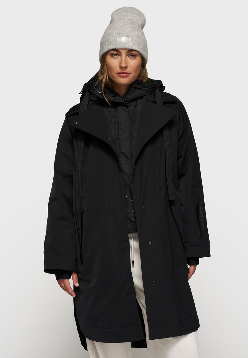 Abrigo largo negro impermeable con un collar alto, diseño de doble botonadura, cordones ajustables y bolsillos laterales. Combinado con un gorro gris.