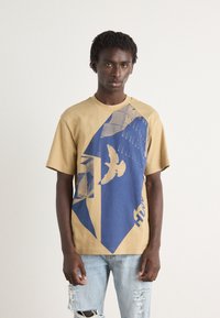 Beige bomull t-skjorte med blå grafisk design som inkluderer en fugl og geometriske mønstre. Har korte ermer og rund hals. Revne lyse denim jeans.