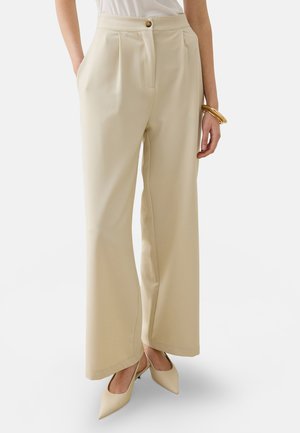 Kvinde iført cremefarvede bukser med højtaljet, vidde ben og sidelommer, kombineret med cremefarvede spidse slingback-hæle og guldarmbånd.