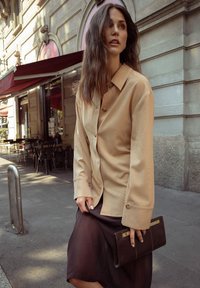 Beige, long-sleeve button-up shirt with a ribbed texture, paired with a dark, flowy skirt and a slim brown clutch with metallic accents.