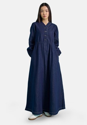Kvinde iført en lang mørkeblå denimkjole med lange ærmer, knapper foran og hvide sneakers, stående med hænderne i lommerne.