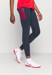 Marinblå träningstights med "BARCA"-logotyp i fet röd, slät textur, slim fit, matchas med vita fotbollsskor med röda accenter.