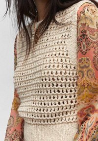 Cremefarbener, gehäkelter Strickpullover mit offenem Muster, der an der Taille eine gerippte Textur aufweist. Kombiniert mit einem gemusterten, mehrfarbigen, durchsichtigen Ärmel.