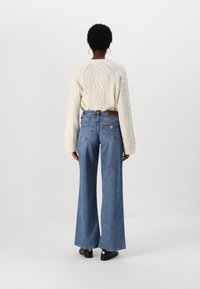 Persona con cabello corto que lleva un suéter crema de punto trenzado, pantalones vaqueros anchos de talle alto en azul, zapatos negros y pendientes de aro dorados, de pie frente a un fondo blanco.