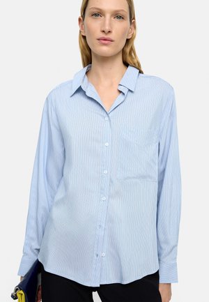 Kobieta w jasnoniebieskiej, długorękawowej koszuli na guziki w cienkie białe paski, trzymająca czarno-żółtą kopertówkę.