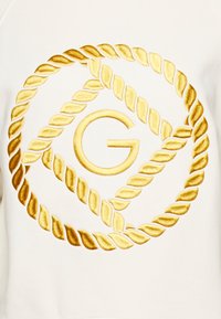 Vit sweatshirt med ett stort guldinbroderat logotypdesign som har repmönster som omger ett centralt "G"-bokstav.