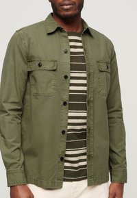 Olive green button-down shirt z kieszeniami na piersi, zestawiony z pasiastą ciemnozieloną i kremową koszulką. Gładki materiał, luźny krój.