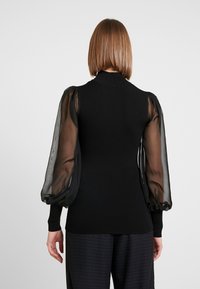 Maglione nero a coste con maniche a sbuffo trasparenti. Collo alto e corpo aderente. Accentuato da un tessuto testurizzato per le maniche e dettagli sui polsini.