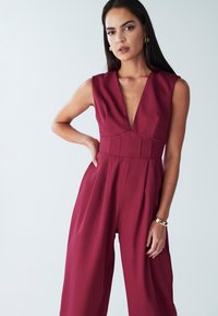 Mono sin mangas color burdeos con un escote en V pronunciado, corpiño plisado y diseño de pierna ancha. Textura de tela suave con detalles mínimos.