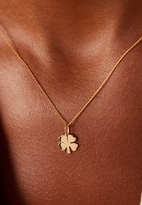 Gouden ketting met een klavertje vier hanger, gladde textuur, met een gepolijste afwerking, rustend tegen een gebruinde huid.