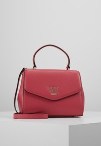 Pieni punainen nahkainen DKNY-käsilaukku, jossa on yläkahva ja irrotettava olkahihna, valkoisella pinnalla harmaata taustaa vasten.