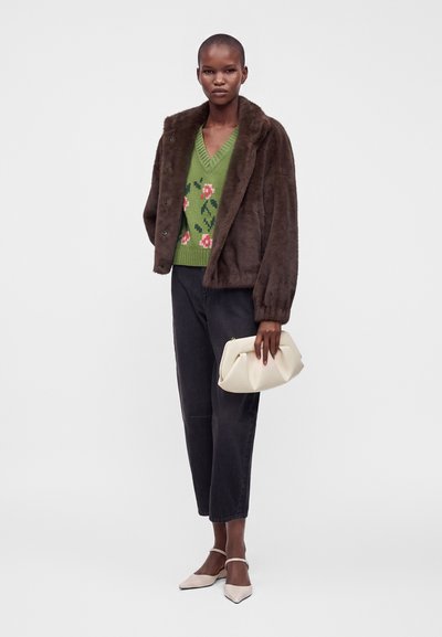 Femme vêtue d'une veste en fourrure marron, d'un pull vert à motifs floraux, d'un pantalon noir, de chaussures blanches pointues, tenant une pochette blanche sur un fond uni.