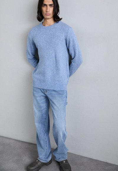 Light blue knitted sweater with a round neckline, paired with faded blue denim jeans and black shoes, standing against a plain backdrop.
