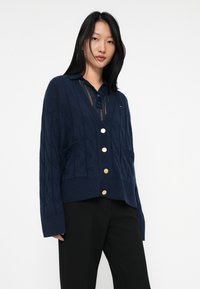 Joven mujer vistiendo un cárdigan de punto trenzado azul marino con botones dorados sobre una camisa de cuello azul marino y pantalones negros, contra un fondo blanco.