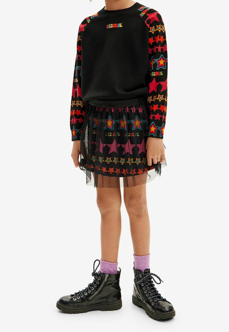 Sweat-shirt noir avec des manches à motifs d'étoiles colorées, jupe en tulle superposée avec imprimés d'étoiles, bottes de combat noires et chaussettes violettes à la cheville.