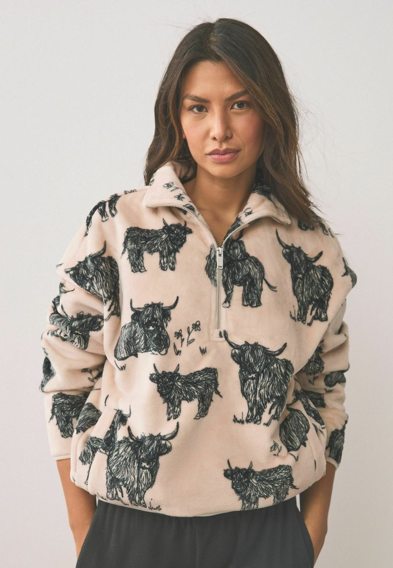 Femme portant un pull en polaire beige orné de croquis noirs de bétail highland, debout les mains dans les poches devant un fond uni.