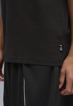 Close-up van het bovenlichaam van een persoon die een zwart shirt en mesh broek draagt, met een klein Puma-logolabel aan de zijnaad van het shirt.