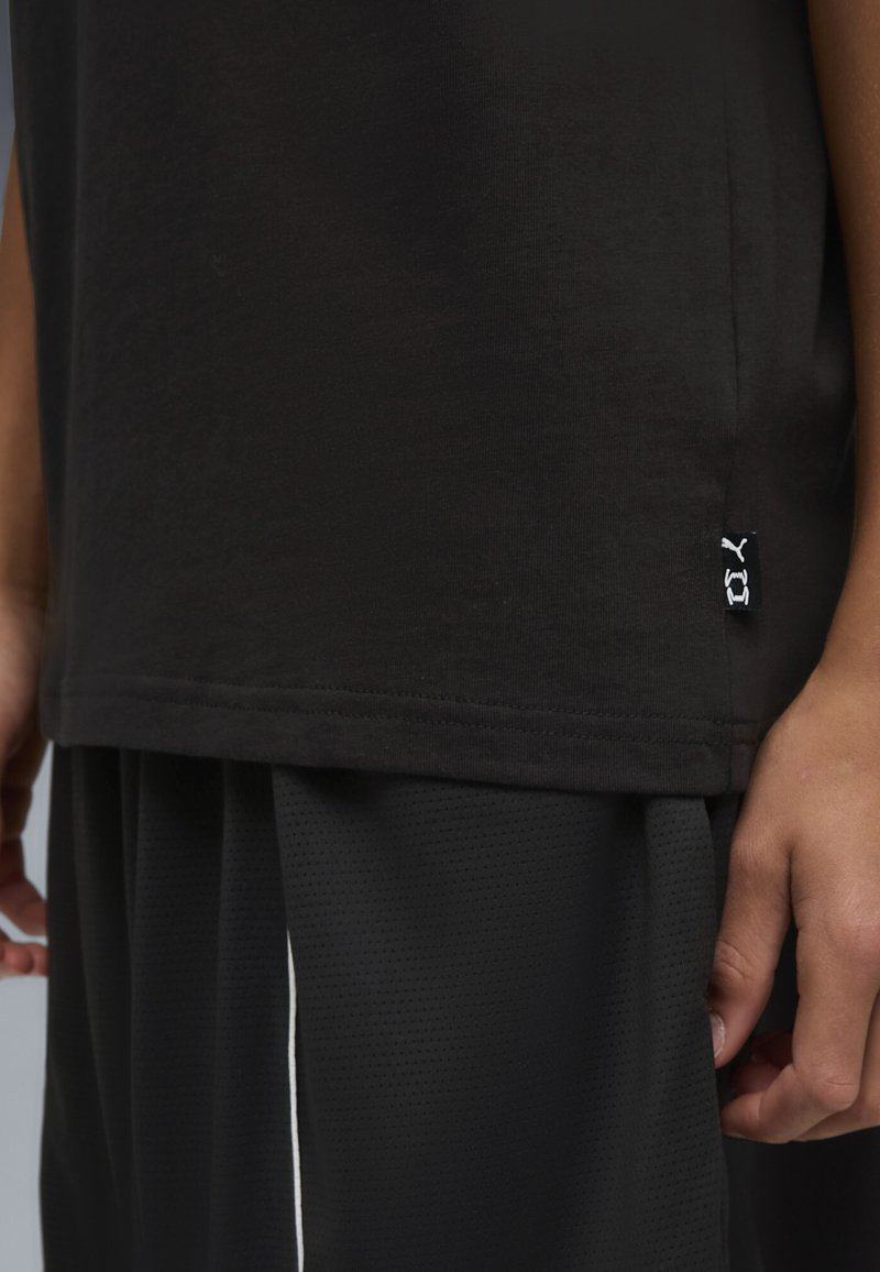 Κοντινό πλάνο του κορμού ενός ατόμου που φοράει μαύρο πουκάμισο και παντελόνι από δίχτυ, με μια μικρή ετικέτα λογότυπου Puma στη ραφή του πουκάμισου στο πλάι.