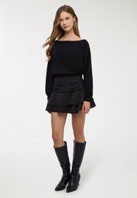 Top nero a maniche lunghe con scollo a barca abbinato a una minigonna nera a balze stratificate e stivali neri con tacco fino al ginocchio.