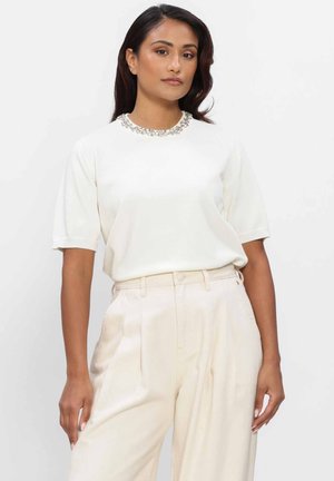 Vrouw die een wit shirt met korte mouwen en een met juwelen versierde halslijn draagt, samen met hooggetailleerde crèmekleurige geplooide broek, staand tegen een witte achtergrond.