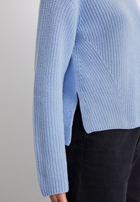 Helltaußer, gestrickter Pullover mit Rippenstruktur, langen Ärmeln, einem seitlichen Schlitz und einer lässigen Passform. Kombiniert mit schwarzen Hosen.