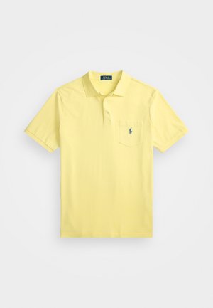 Lyhythihainen polo-paita vaaleankeltaisesta puuvillasta, jossa on kolmesta napista muodostuva kaulus ja vasemmalla rintapoiminta, jossa on pieni sininen logo.
