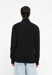 Svart ribbestrikket genser med høy krage og lange ermer, kombinert med lyseblå denimbukser. Enkel design, teksturert stoff.