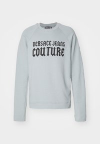 Světle šedá mikina s černým textem "VERSACE JEANS COUTURE". Má kulatý výstřih a žebrované manžety s volnějším střihem.