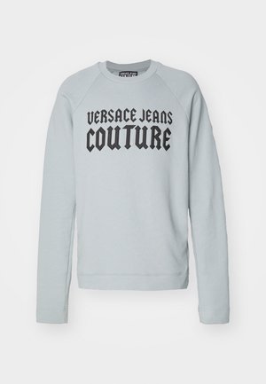 Svetlosivý mikina s čiernym textom "VERSACE JEANS COUTURE". Obsahuje okrúhly výstrih a rebrované manžety s uvoľneným strihom.