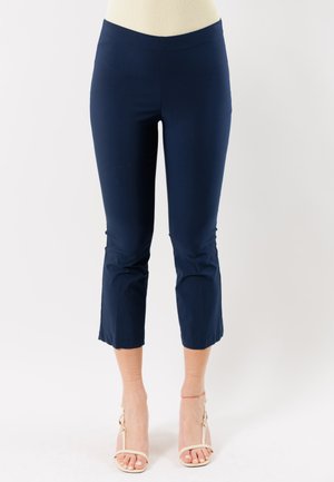 Donna che indossa pantaloni corti blu navy e sandali beige con tacchi alti, in piedi contro uno sfondo bianco uniforme.