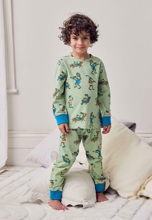 Bambino con capelli ricci in piedi a piedi nudi su cuscini, indossa un pigiama verde con divertenti illustrazioni di rane e polsini blu.