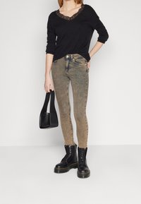 Top negro con escote de encaje, jeans ajustados beige y botas de combate negras. Sostiene un pequeño bolso negro, el fondo es blanco liso.