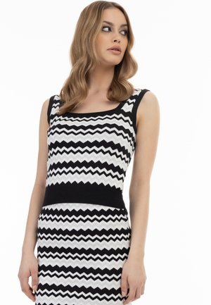 Femeie cu păr blond ondulat, purtând o rochie fără mâneci cu model zigzag alb-negru, privind spre stânga pe un fundal alb.