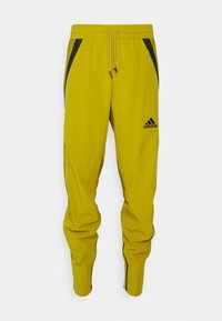 Trainingsanzughose in leuchtendem Gelb, aus leichtem Material gefertigt. Mit schwarzen Seitentaschen und Reißverschlüssen an den Knöcheln. Adidas-Logo sichtbar.