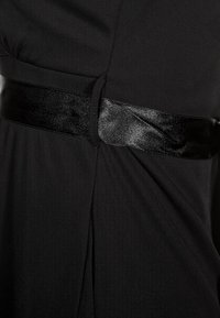 Musta mekko, jossa on satiininen musta vyö, sivuhalkiot ja pehmeä rakenne. Kangas vaikuttaa sileältä ja siinä on hieman kiiltoa.