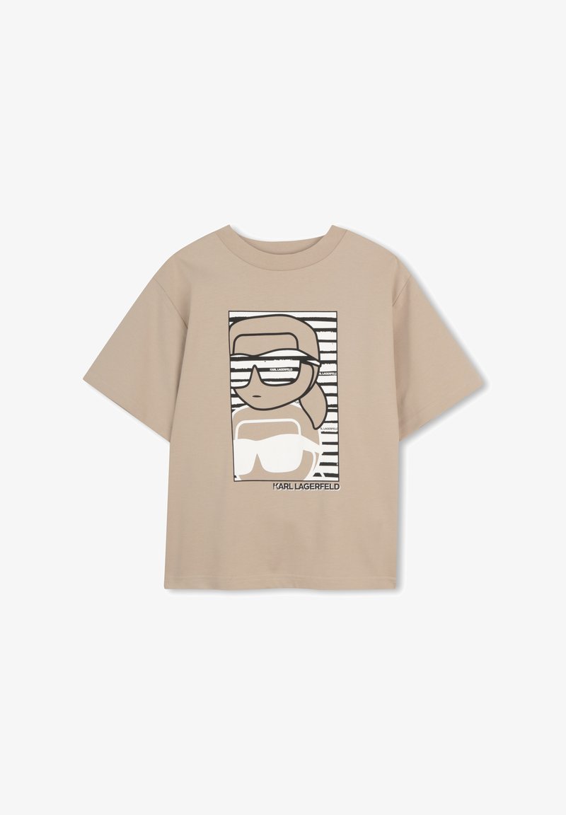 Μπεζ κοντομάνικη μπλούζα με καρτούν γραφικό που απεικονίζει άτομο με γυαλιά ηλίου και αλογοουρά, με το κείμενο «KARL LAGERFELD» κάτω από το σχέδιο.