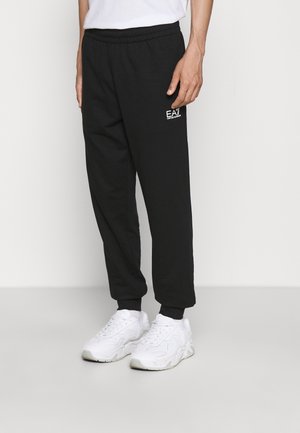 Mann trägt schwarze Jogginghosen mit weißem EA7 Emporio Armani-Logo und weiße Sneakers, steht vor einem schlichten hellen Hintergrund.