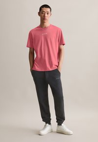 Roze katoenen t-shirt met een ronde hals en geborduurde tekst, gecombineerd met zwarte joggingbroek en witte sneakers, gesitueerd tegen een neutrale achtergrond.