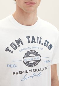 White cotton T-shirt featuring blue and black graphic print with brand name, text details, and circular logo. Round neckline, standard fit.