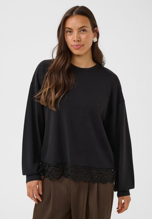 Femme aux longs cheveux bruns portant un sweat noir ample avec une bordure en dentelle et un pantalon marron à jambes larges, debout devant un fond clair uni.