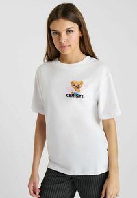 T-shirt en coton blanc avec un col rond, présentant un graphique d'un ours portant des lunettes de soleil et le texte "CERISES" en noir. Manches courtes.