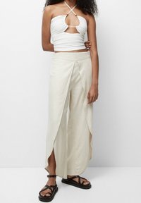 Un crop top blanc texturé avec une découpe à l'avant et un col halter, assorti à un pantalon large crème avec des fentes latérales. Sandales noires à brides.