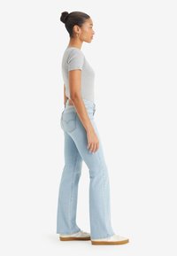 Femme avec les cheveux en chignon, portant des boucles d'oreilles en or, un haut gris ajusté à manches courtes, un jean bootcut bleu clair et des baskets blanches, se tenant de profil.