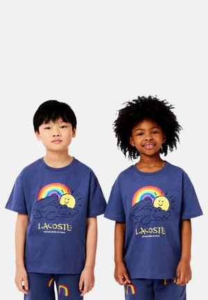 Zwei Kinder in passenden navyblauen Lacoste-Outfits mit Regenbogen-, Sonnen- und Wolkengrafiken auf T-Shirts und Hosen, die vor einem weißen Hintergrund stehen.